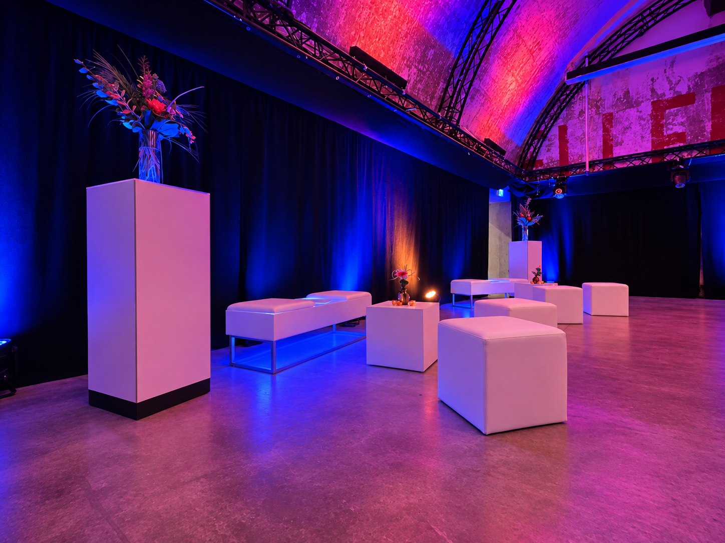 Lounge mit weißen Sitzbänken, Hockern und Stelen im blau-violett beleuchteten Gewölbe